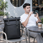 Le Nomade, sac à dos ordinateur Roll Top noir posé à côté d’un utilisateur au café